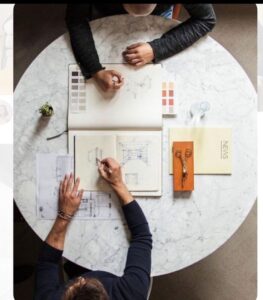 Top-down view of two people sketching architectural designs on a round marble table with color swatches, blueprints, and stationery.
